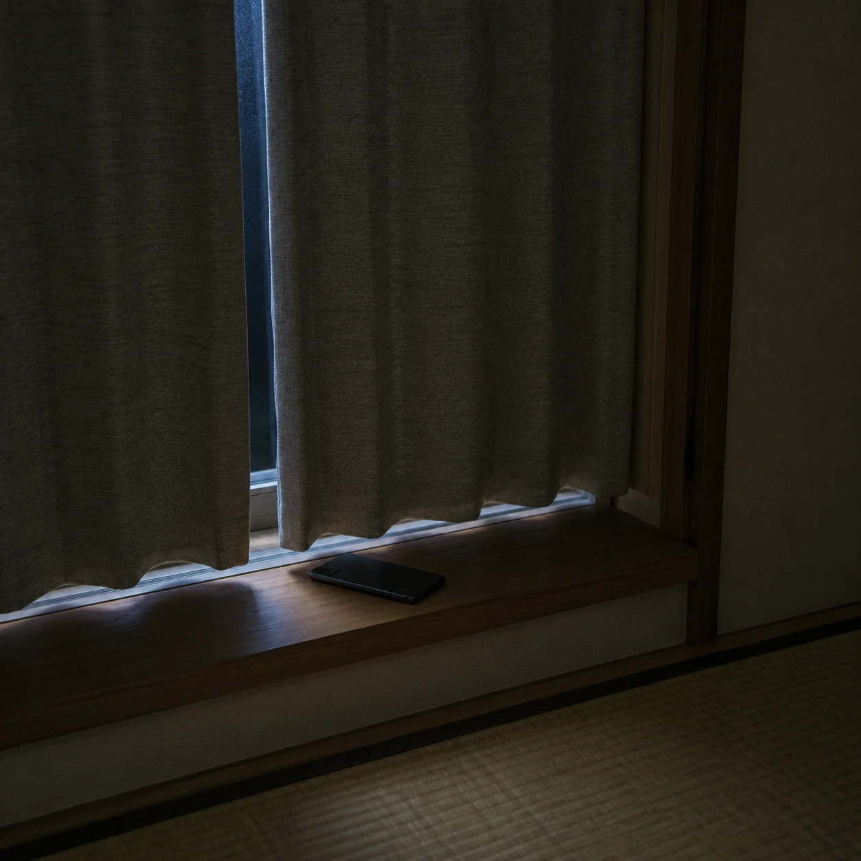 夜の和室の窓辺。閉じたカーテンのすきまから青白い光が差し込み、木の窓台に画面を伏せたスマートフォンが一台だけ置かれている